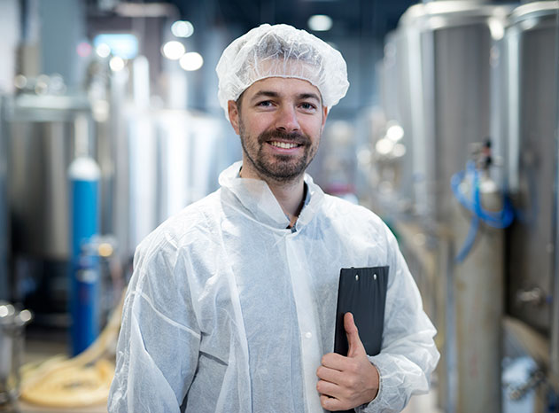 Técnico con bata blanca y gorro de higiene sonriendo con una carpeta en una planta de producción alimentaria